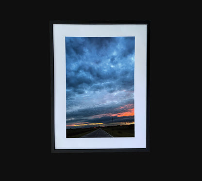 Fine art print of large clouds after a storm and sunset over rural road.  Framed view
